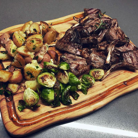A large freeform wood carving board with a deep groove or juice rail catching spills from the lamb cutlets, loaded with roast potatoes and brussel sprouts, made designed by Nature's Boards.