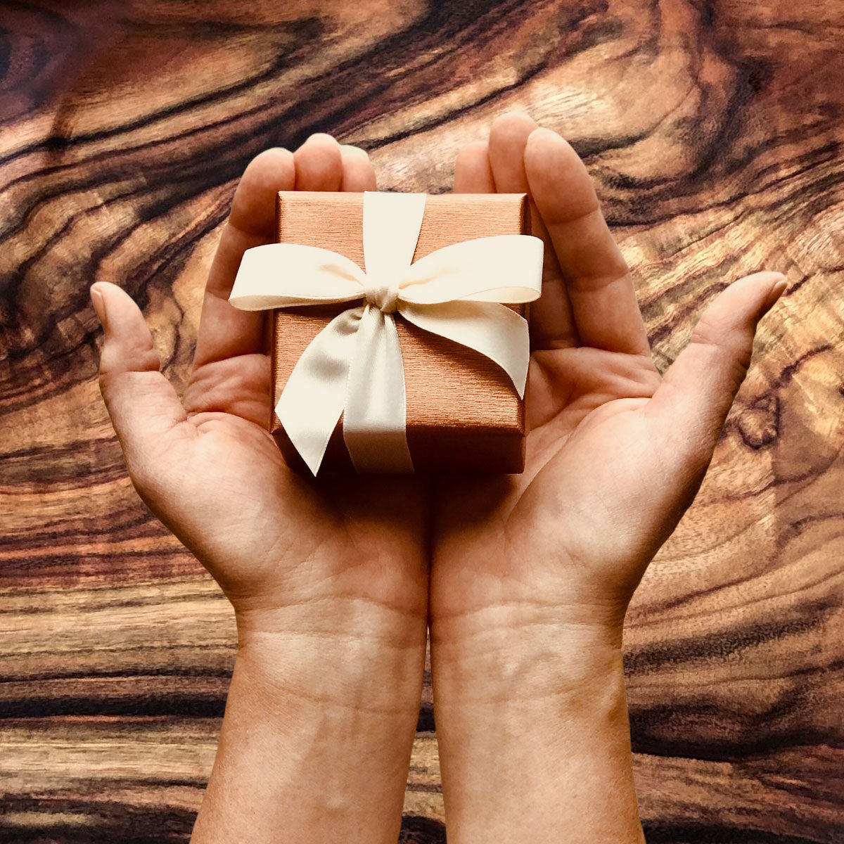 Two hands holding a small wrapped gift box with a bow, with beautiful camphor laurel timber as the background. 